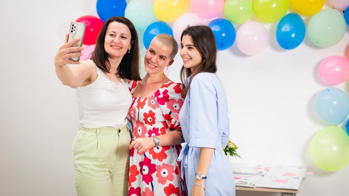 Three women joyfully taking a selfie against a colorful balloon backdrop indoors.